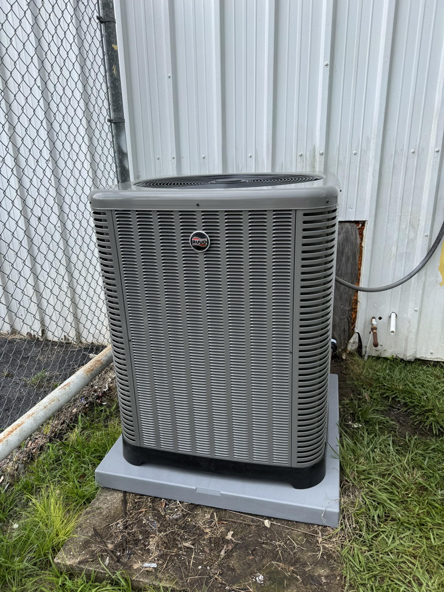 Ruud outdoor air conditioning condenser unit installed on a pad beside a building and chain-link fence