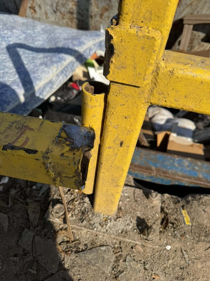 Worn yellow metal cross with chipped paint and burn damage, standing among debris and rubble outdoors