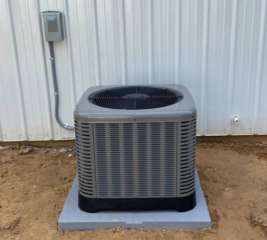 Outdoor AC condenser unit on a concrete pad beside a metal building with an electrical disconnect box mounted on the wall
