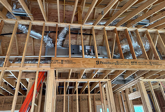 Exposed wooden framing of a house under construction, showing HVAC ductwork and an air handler unit in the ceiling