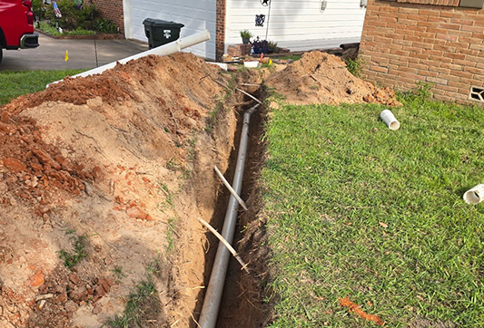 Deep trench dug in a residential front yard with a gray pipe laid along the bottom, surrounded by excavated red clay soil